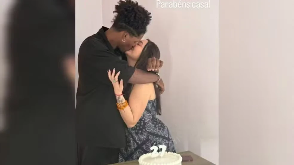 Casal celebra o aniversário com um beijo ao lado de um bolo decorado com o número 27, simbolizando a idade compartilhada.