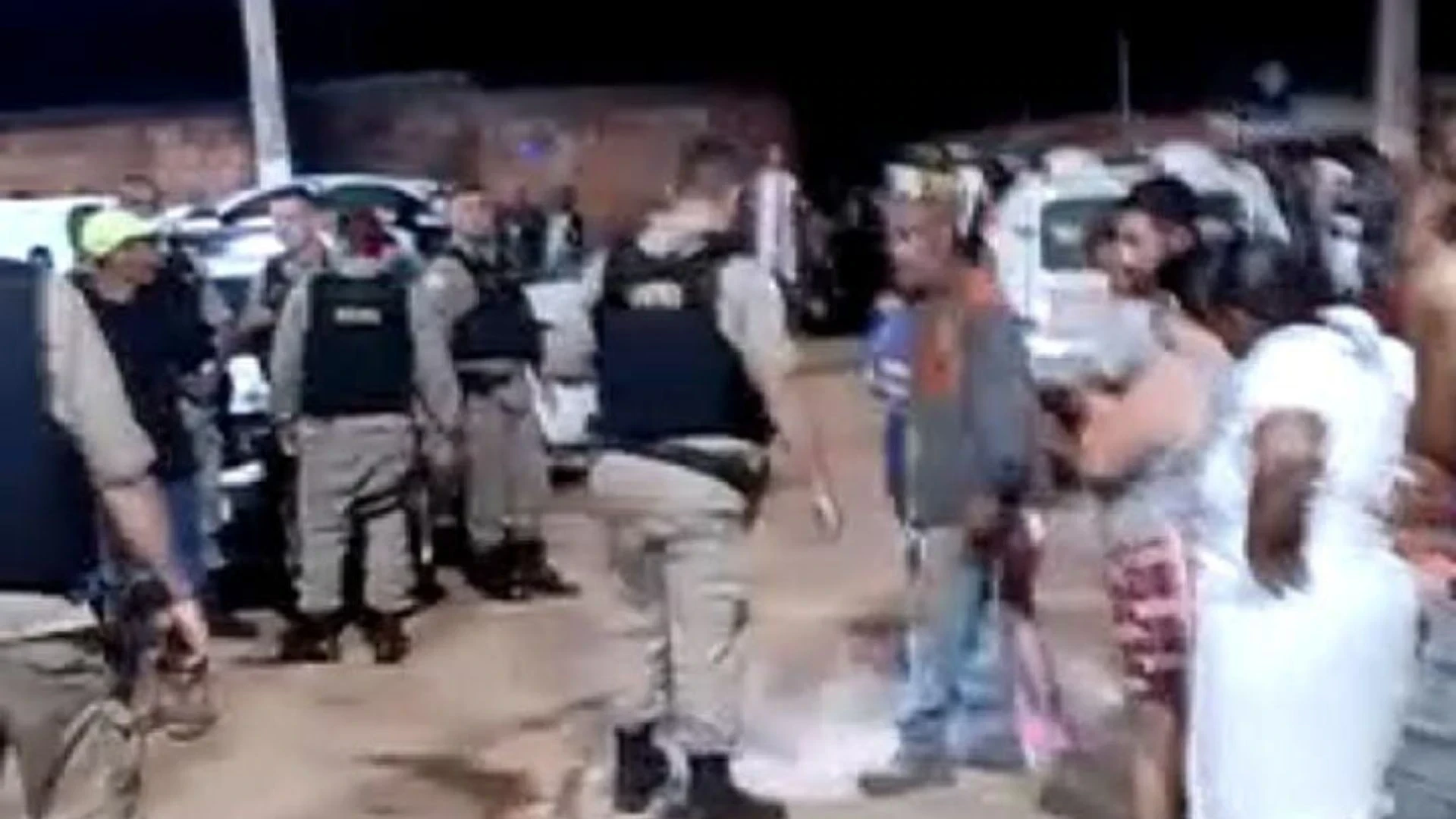 Policiais interagem com pessoas em uma área urbana à noite, reforçando a segurança local. As circunstâncias envolvidas não estão claras.