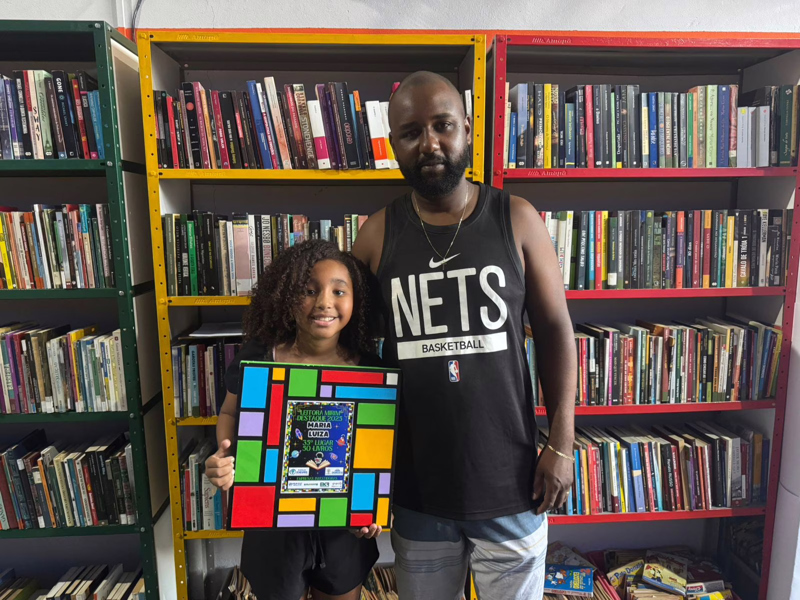 Anderson da Silva posa ao lado da filha, Maria Luísa Rodrigues, uma das leitoras mirins premiadas pelo projeto do Instituto IDEIAS. A imagem simboliza o apoio da família à iniciativa que incentiva o hábito da leitura desde a infância e reconhece o esforço das crianças que se destacaram ao longo do ano.